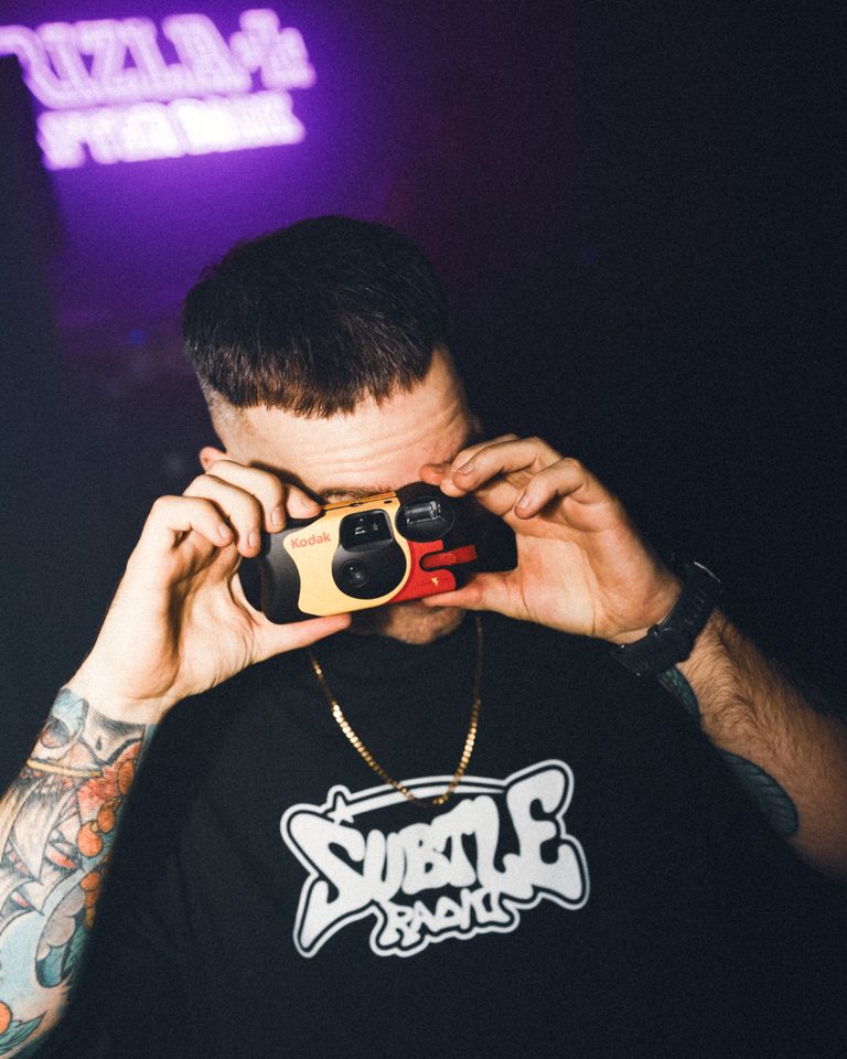 Person holding Kodak camera, neon sign in background, tattooed arm, black t-shirt.