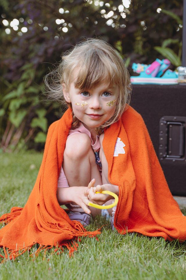 A young child sits on grass wrapped in an orange blanket, holding a yellow object.