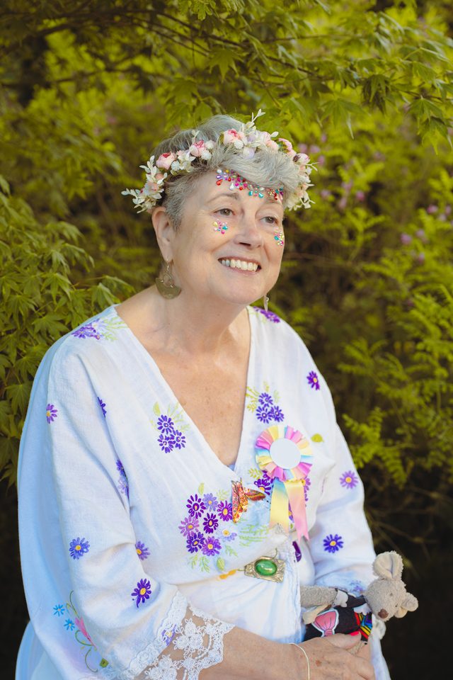 Person in floral attire and crown, smiling outdoors with greenery behind.