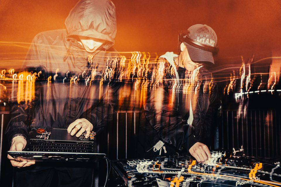 Two DJs at a mixing console under stage lighting, one with laptop, one with headphones.