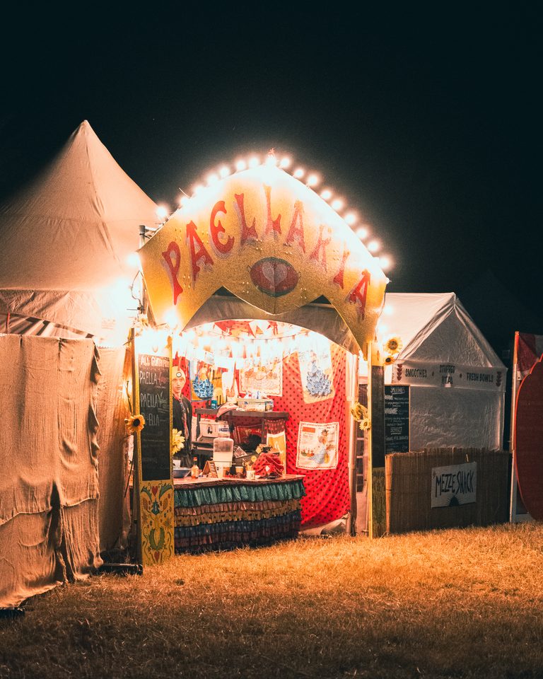 A brightly lit paella stall at night, surrounded by other tents on grass.