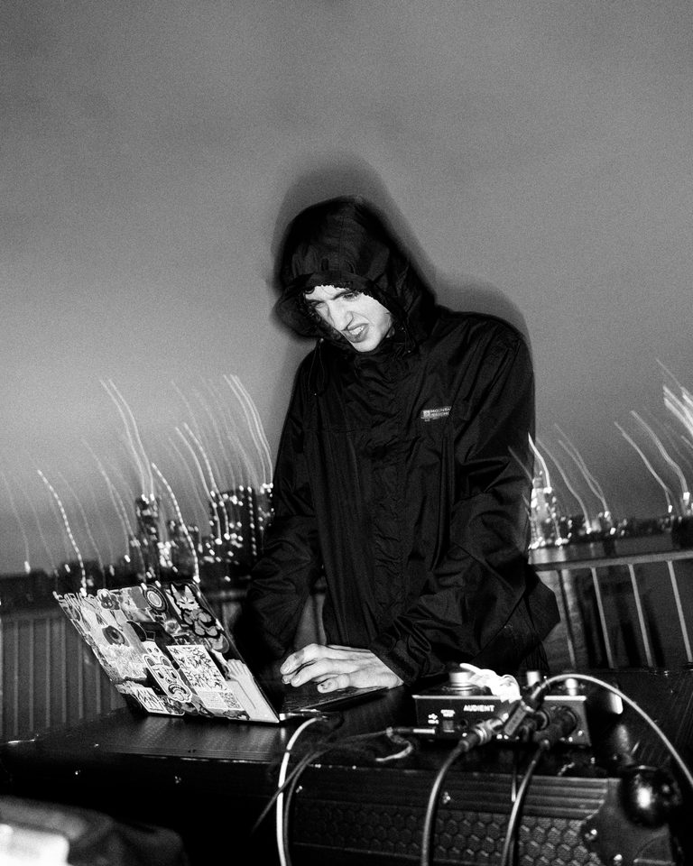 A person in a hooded jacket uses a laptop at a DJ booth with city lights in the background.