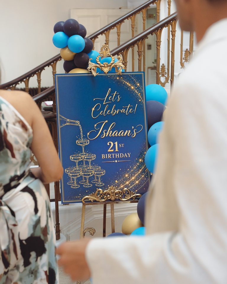 Blue and gold birthday sign for Ishaan's 21st, surrounded by balloons, with guests in foreground.