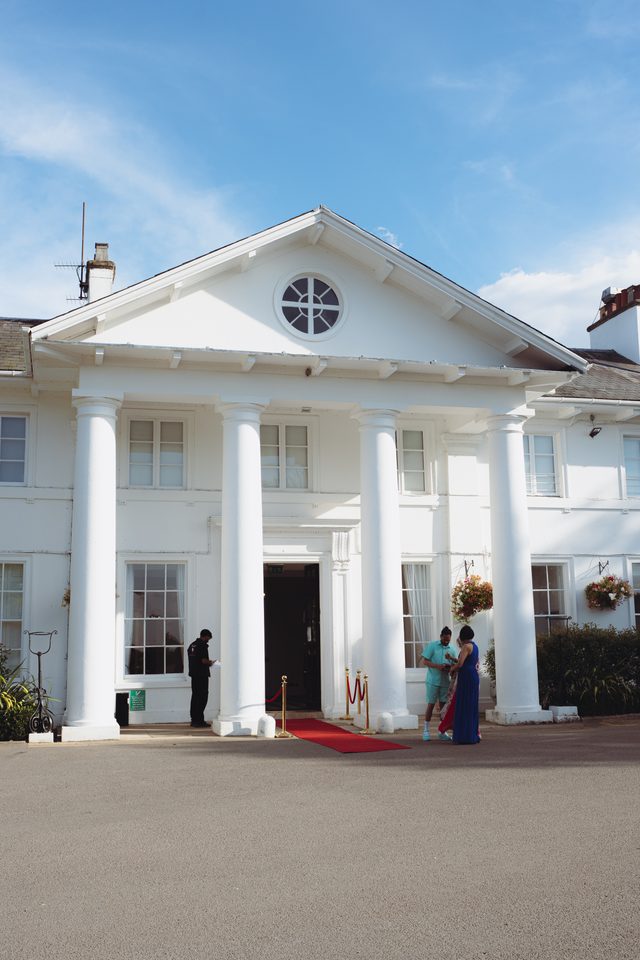 White-columned building with red carpet, two guests conversing near entrance, clear blue sky.