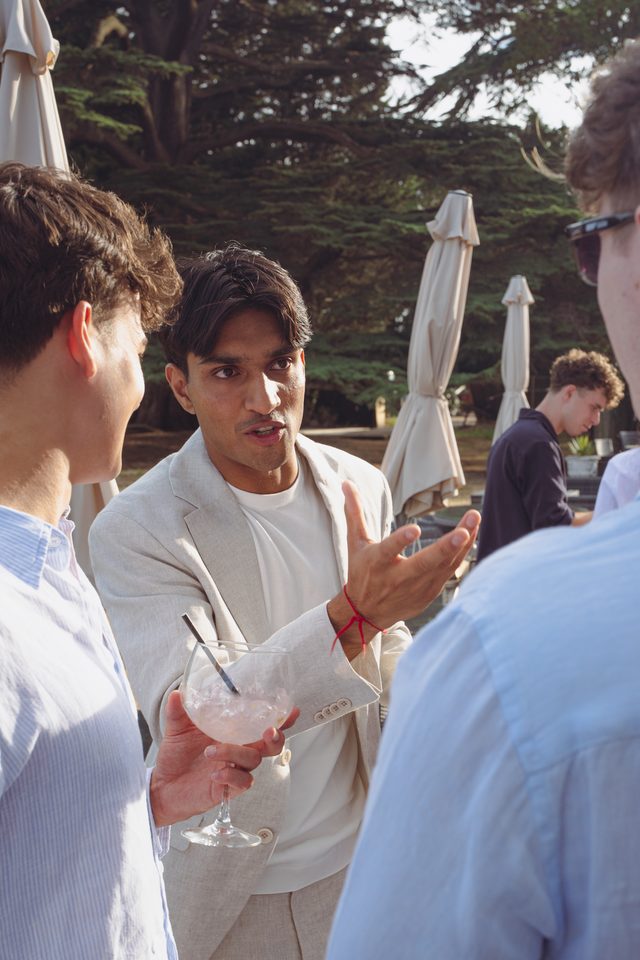 Three men conversing outdoors, one gesturing with a drink in hand.