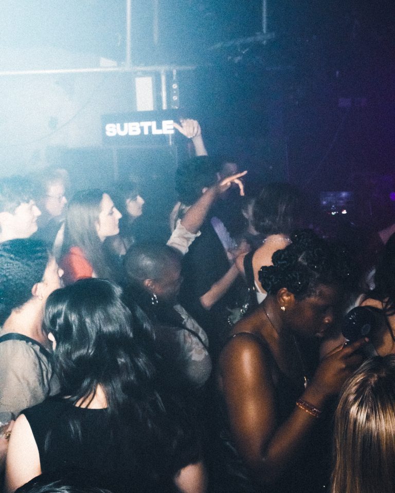 People dancing in a dimly lit club with a 'SUBTLE' sign overhead.