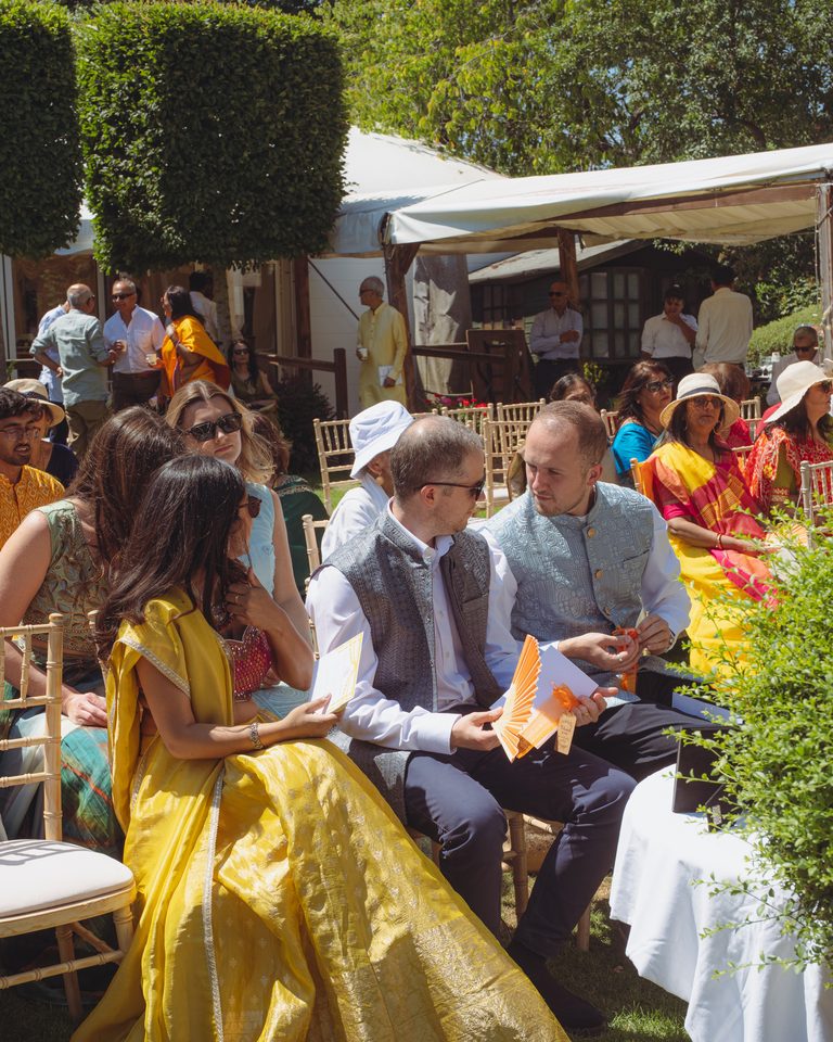 Guests seated outdoors under trees, wearing colorful attire, holding paper items.
