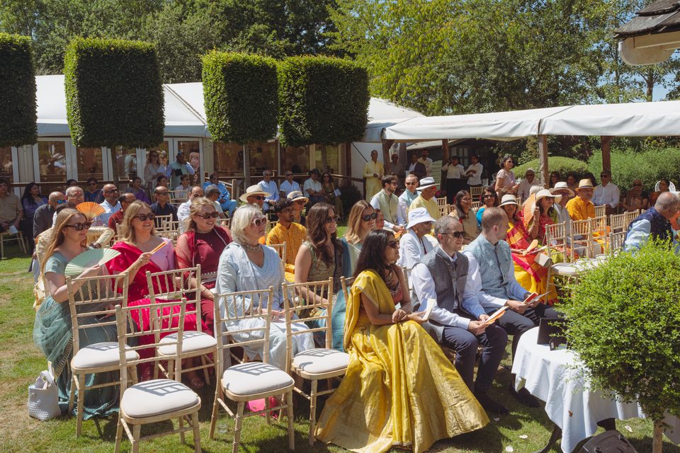 Guests seated outdoors under trees and tents, wearing colorful attire.