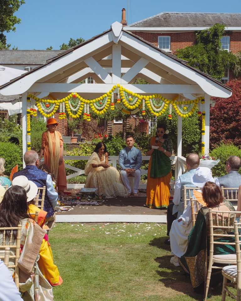 A couple seated under a floral arch with guests watching, sunlit outdoor setting.