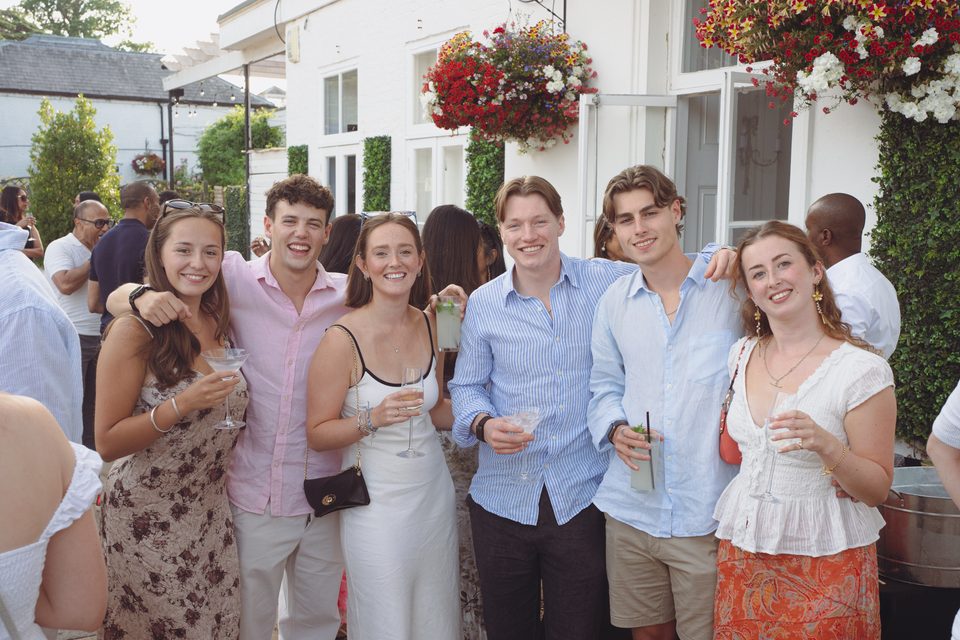 Six people posing together at an outdoor event, holding drinks, with a white building and hanging flowers behind them.