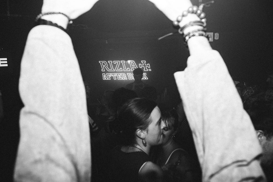Two arms frame a crowd under neon sign, dark indoor club lighting.