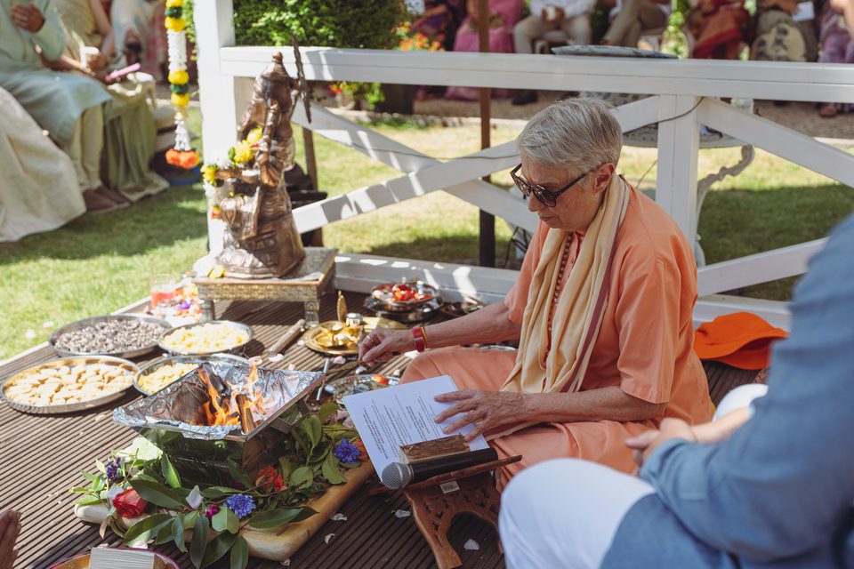 An elder in orange robes reads from a script beside a ceremonial fire and offerings.