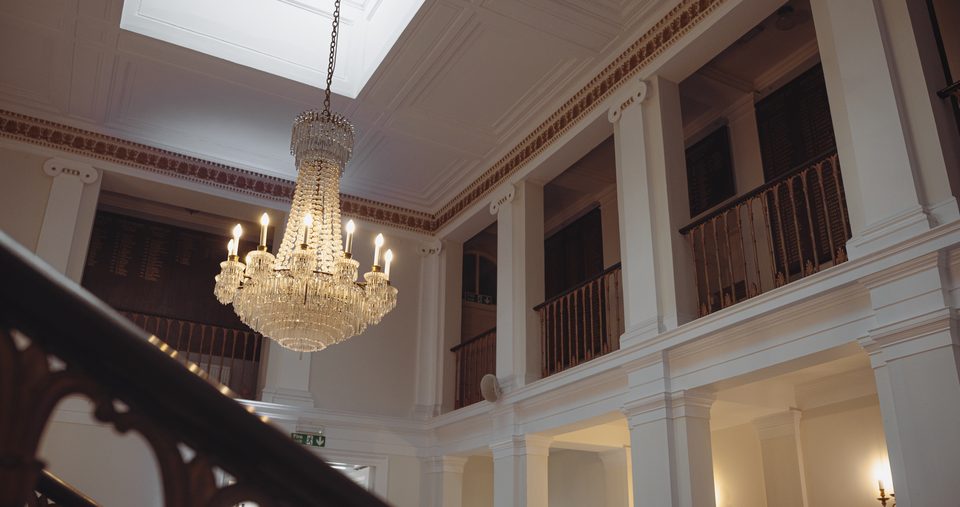 A large crystal chandelier hangs from a high ceiling in a grand indoor space with white columns and ornate railings.
