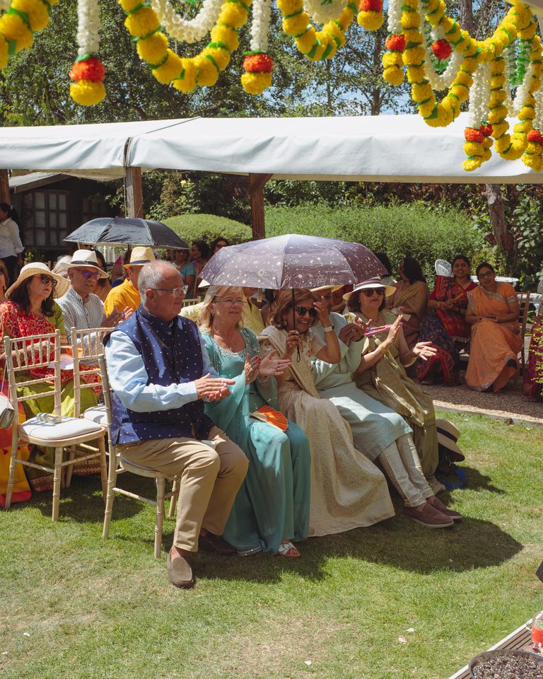 Guests seated under a canopy with floral decorations, clapping in daylight.