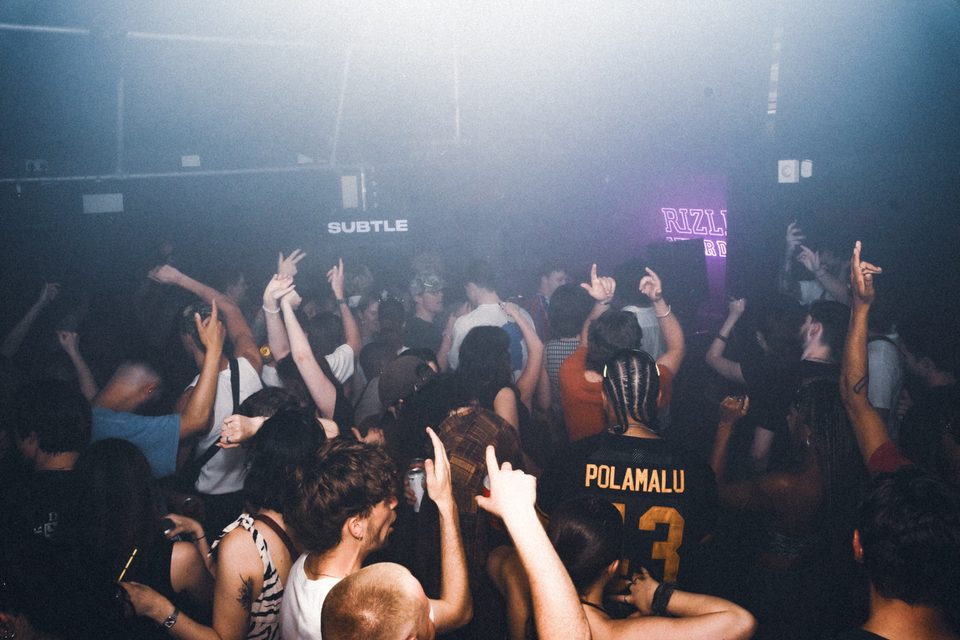Dense crowd at a club with raised hands, illuminated by haze and stage lights.
