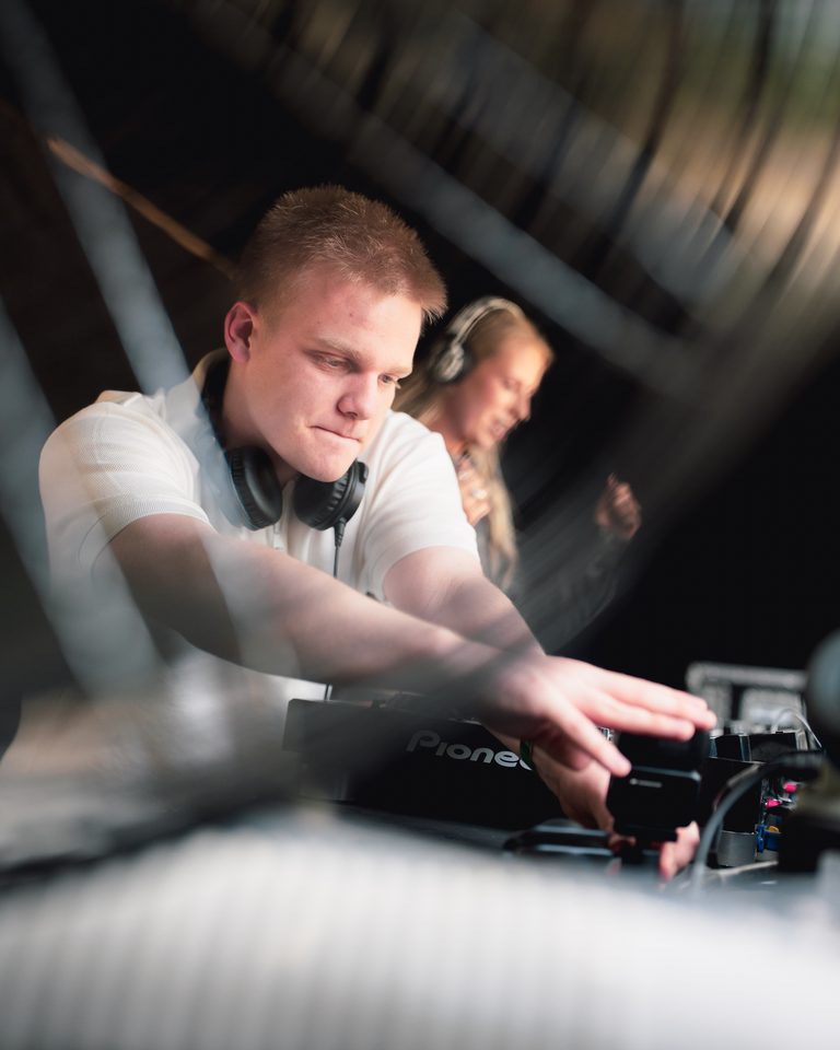 A DJ with headphones adjusting equipment in a dimly lit booth.