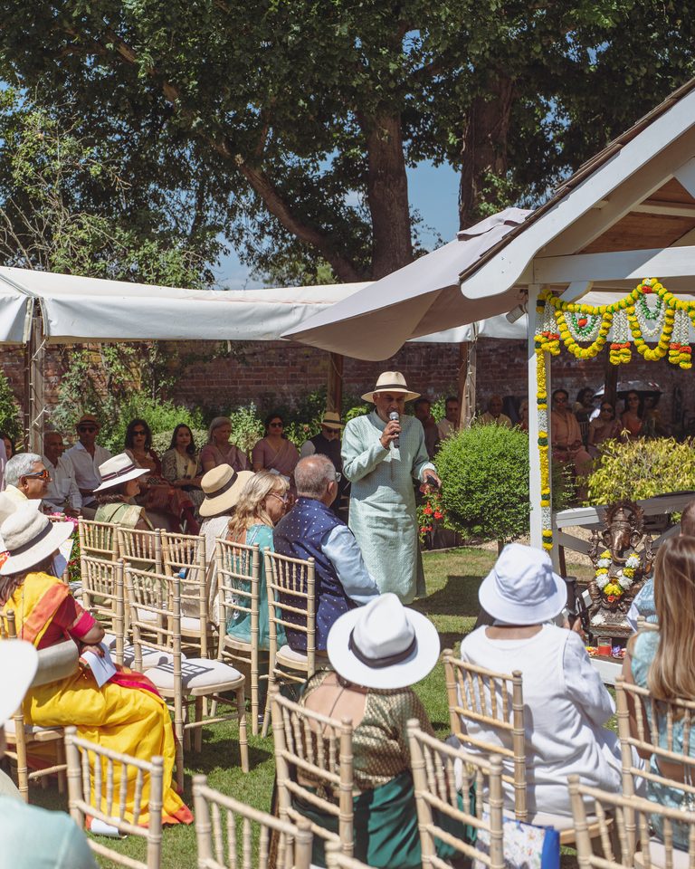 Person speaks into microphone under canopy, guests seated in chairs, trees and sunlit garden background.