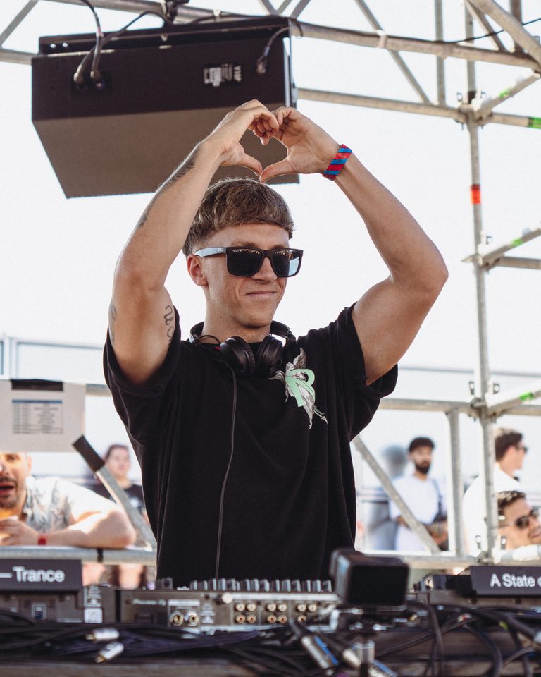 DJ making heart gesture with hands, wearing sunglasses and headphones, standing behind equipment under bright daylight.