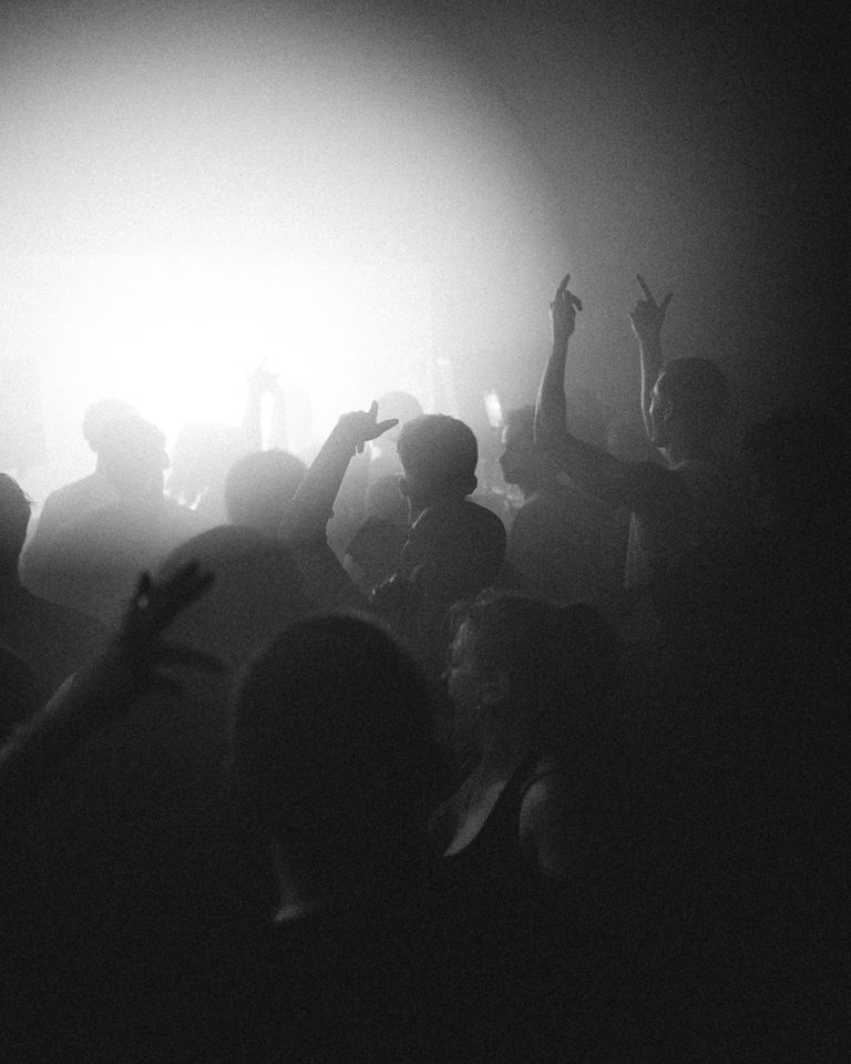 Silhouetted crowd with raised hands under bright stage lighting in a dim club environment.