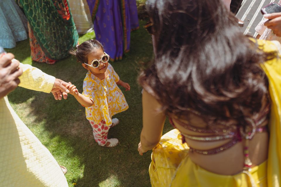 Little girl in yellow outfit and sunglasses, holding hands with adults on grass.