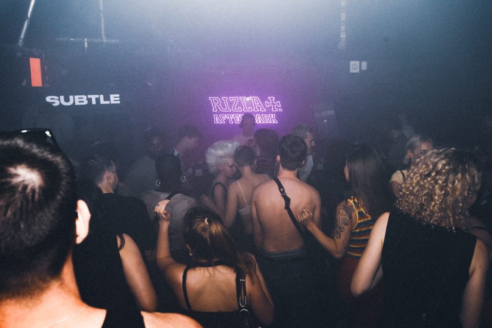 Crowd dancing in a smoky club with purple neon sign.
