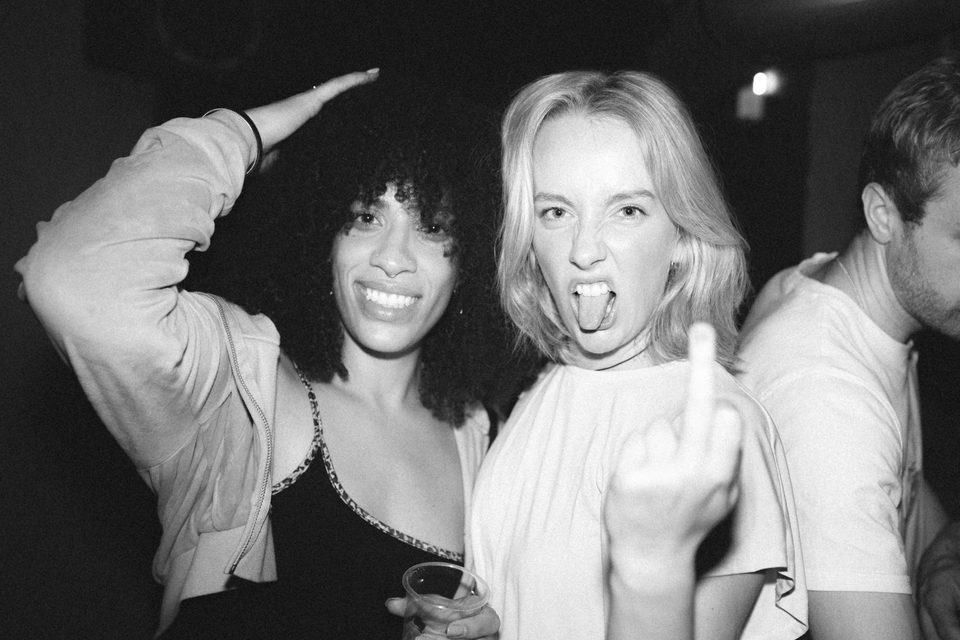 Two women at a nightclub, one making a gesture, the other smiling, in low-light indoor setting.