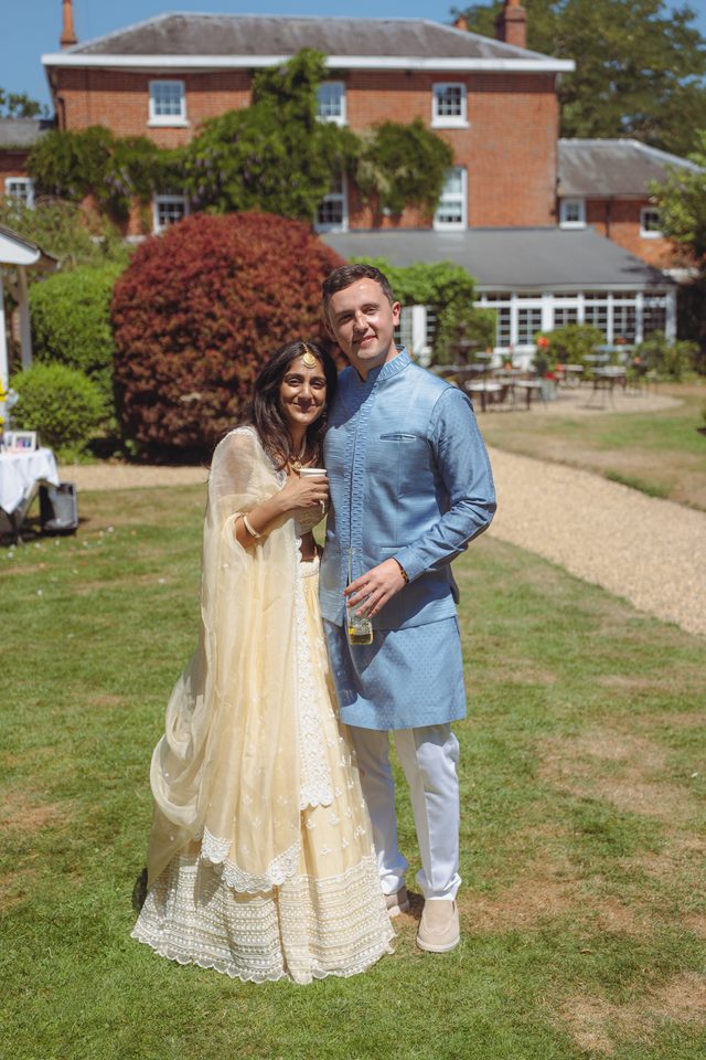 A couple in formal attire standing on grass, holding drinks, with a brick house and garden in background.