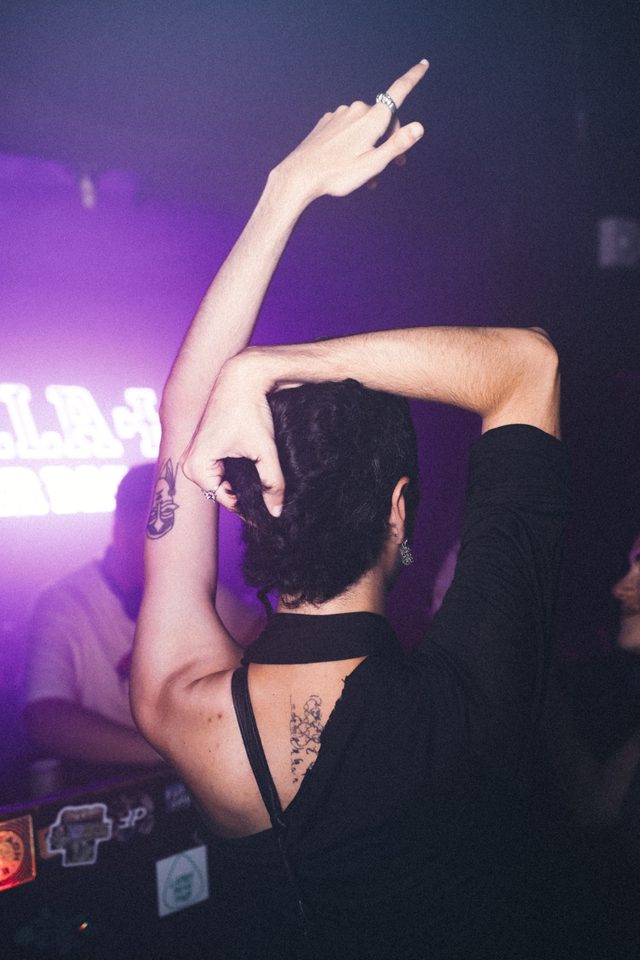 Person dancing with arms raised under purple stage lights in a crowded indoor club.