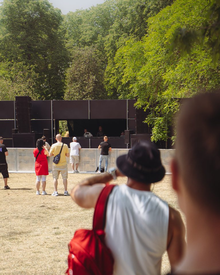 Audience watches performers at a sunlit outdoor stage surrounded by trees.