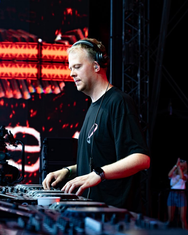 A DJ wearing headphones and a black shirt, hands on equipment, under red stage lighting.