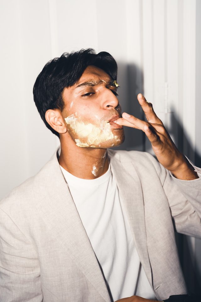 Person with cream on face, hand near mouth, wearing light jacket and white tee.
