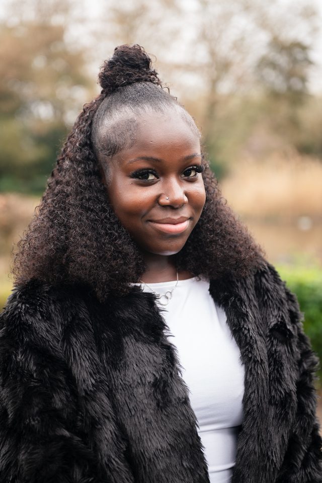 A person in a black fur coat and white top, standing outdoors with blurred trees behind.