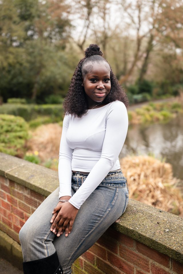 A person sits on a brick wall beside a river, wearing a white top and jeans.