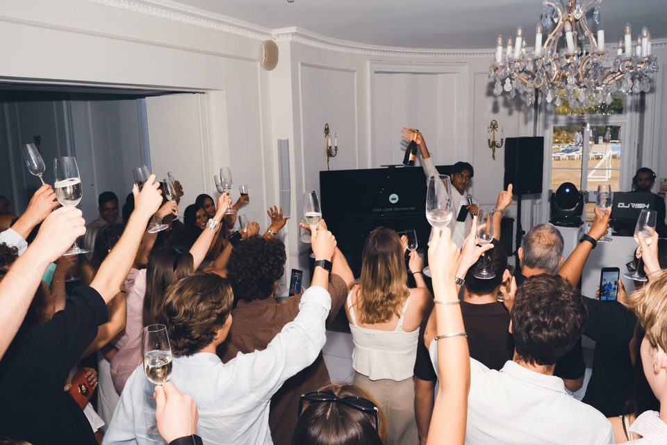People raise glasses in a crowded indoor club, a DJ stands with a bottle, stage lights and a chandelier illuminate the scene.