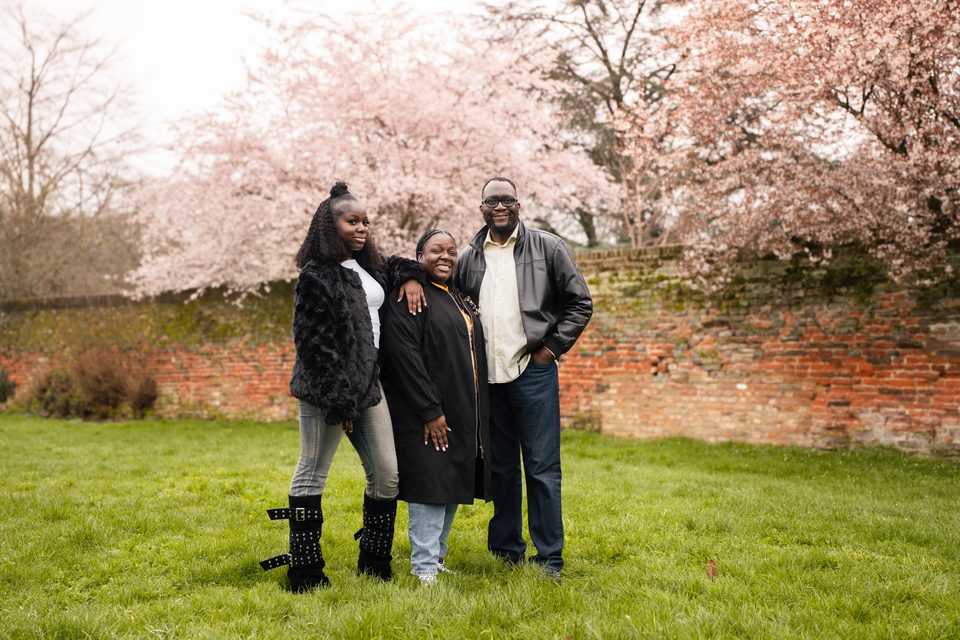Three people standing on grass in front of a brick wall with cherry blossoms.