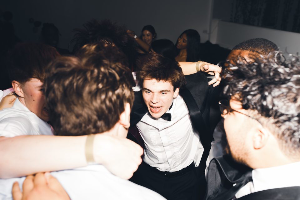Group of young people in formal attire dancing closely together in a dimly lit indoor venue.