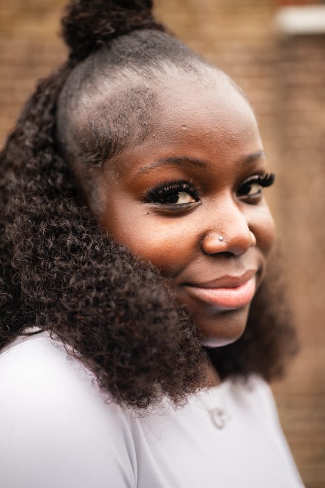 A person with curly hair and nose piercing, wearing a white top, against a blurred brick wall.