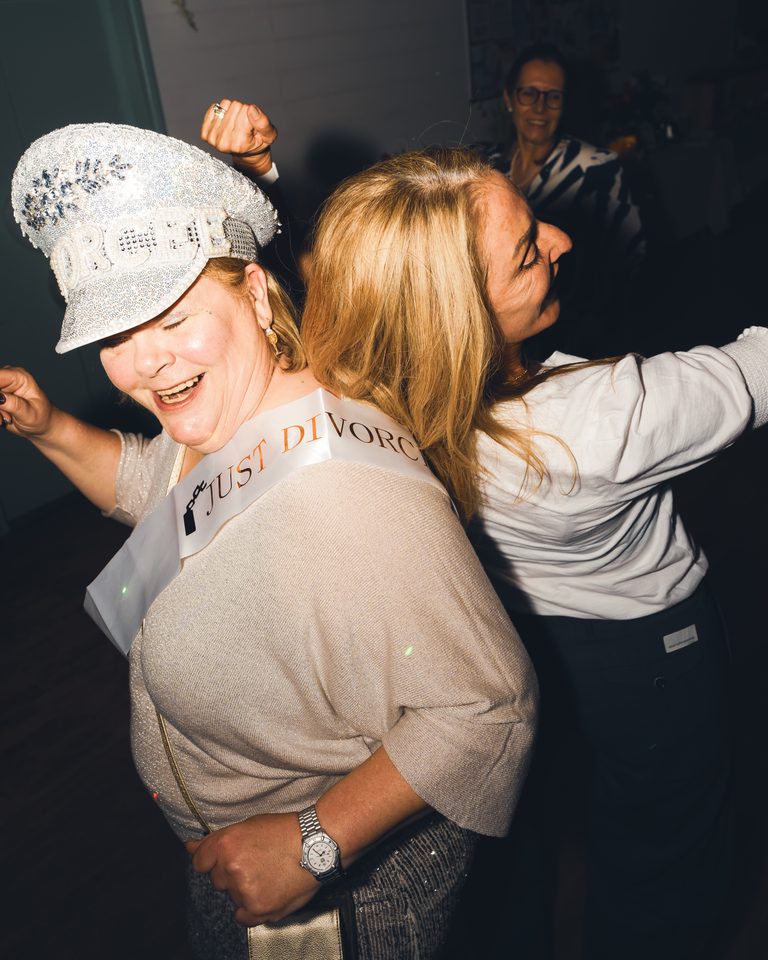 Two women dancing in a dimly lit indoor space, one wearing a 'Just Divorced' sash.