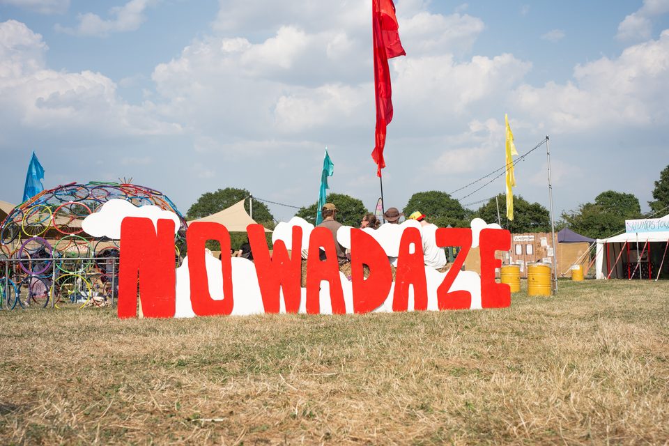 Large red letters spelling 'NOWADAZE' on grass with people and flags in background.