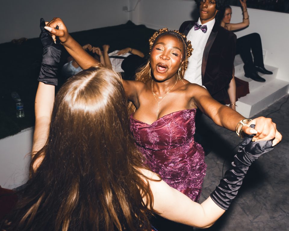 Two women dancing in a dimly lit club, one in a purple dress and gloves.