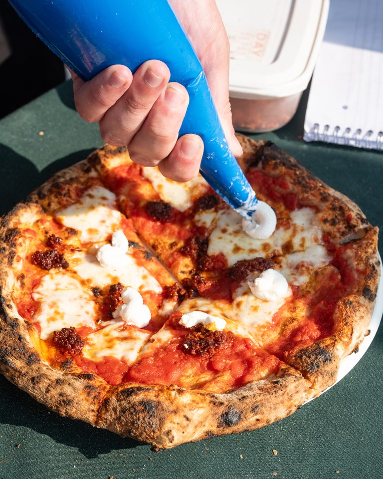 Hand squeezing blue piping bag to add cheese to a charred pizza crust.