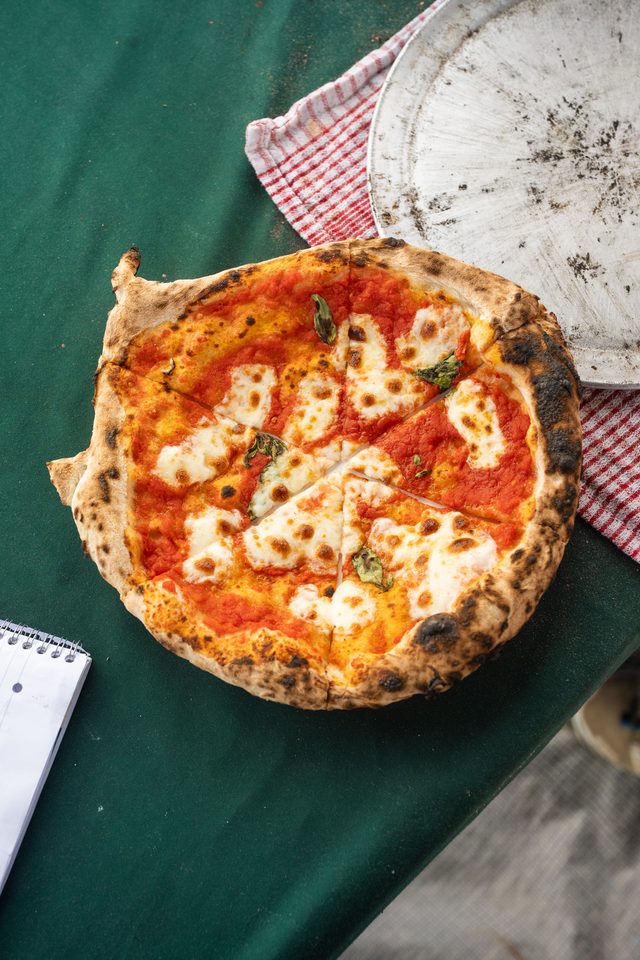 A freshly baked pizza with charred crust sits on a green table beside a notebook and a worn tray.