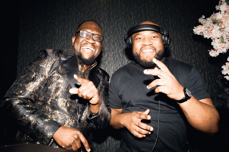 Two men in DJ booth, one with headphones, one in patterned jacket, both smiling and gesturing.
