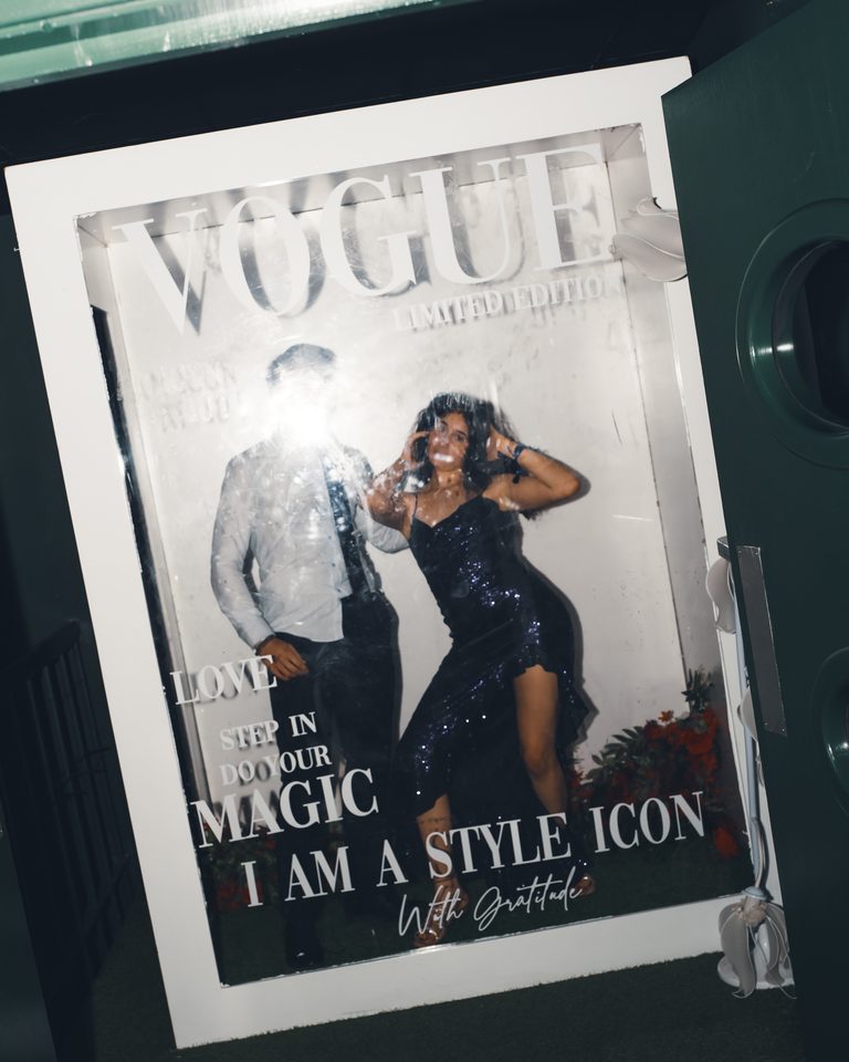 Two people posing in a photo booth with Vogue magazine design, one in sequined dress, one in formal attire.