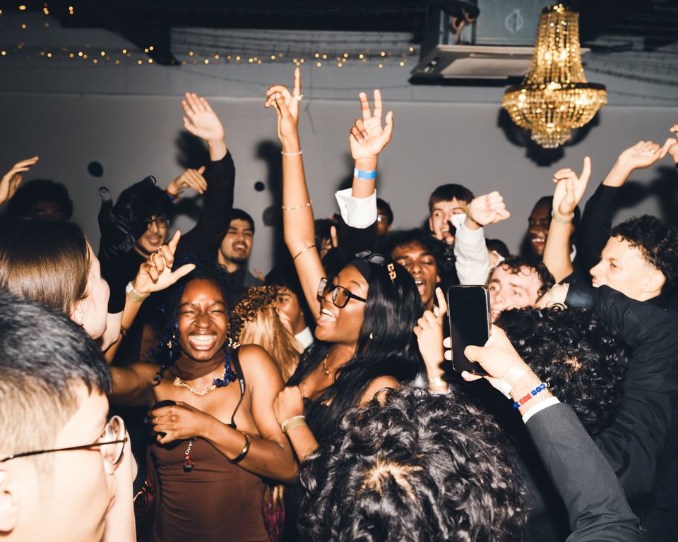People dancing in a crowded indoor club with raised hands and a chandelier overhead.
