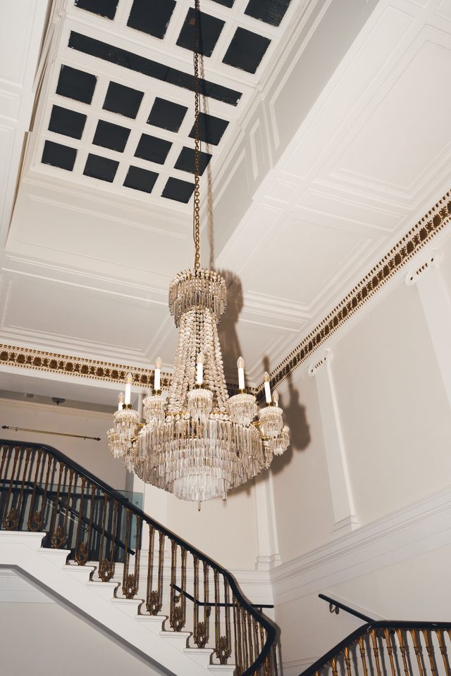 A large crystal chandelier hangs above a white staircase with ornate railings in a grand indoor space.