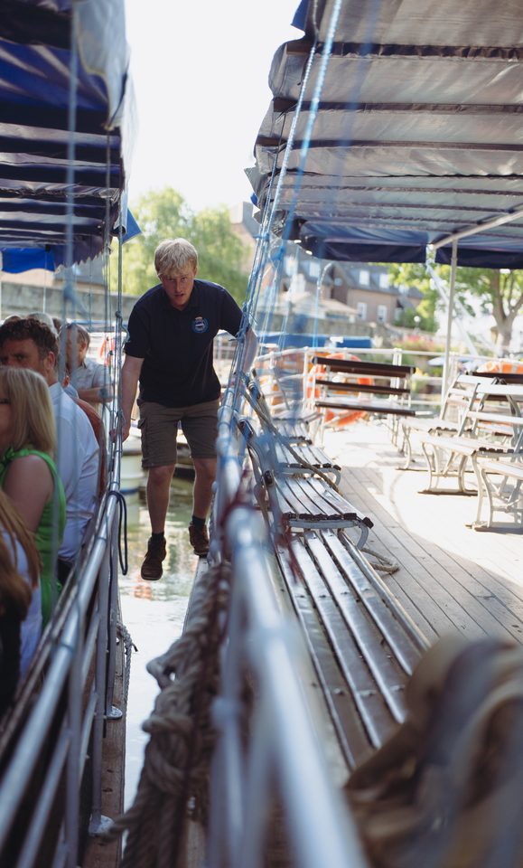 Person in dark shirt walking on boat deck with passengers nearby.