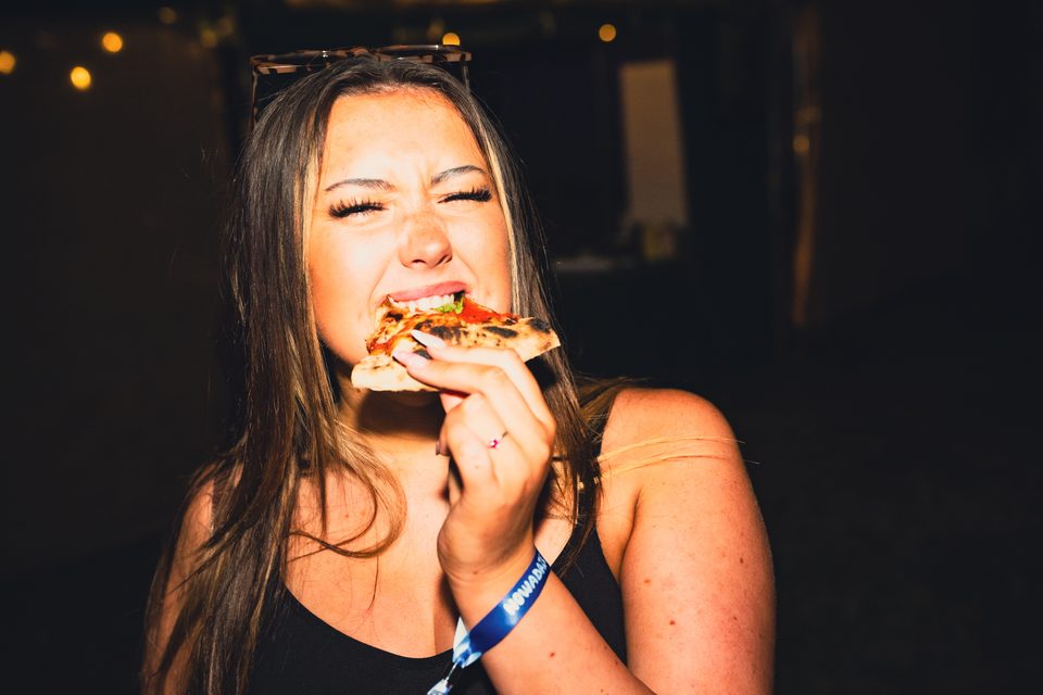 Person eating pizza slice with sunglasses on head, wearing event wristband.