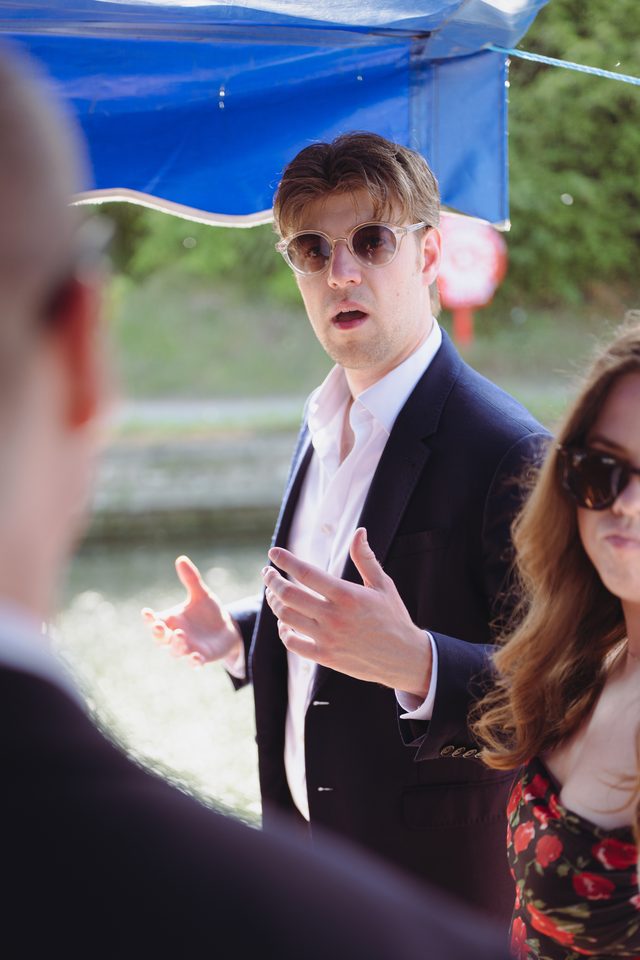 Person in sunglasses and suit gestures while speaking to others under a blue tent.