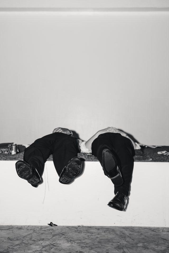 Two individuals in dark attire with feet up, resting on a ledge against a plain wall.